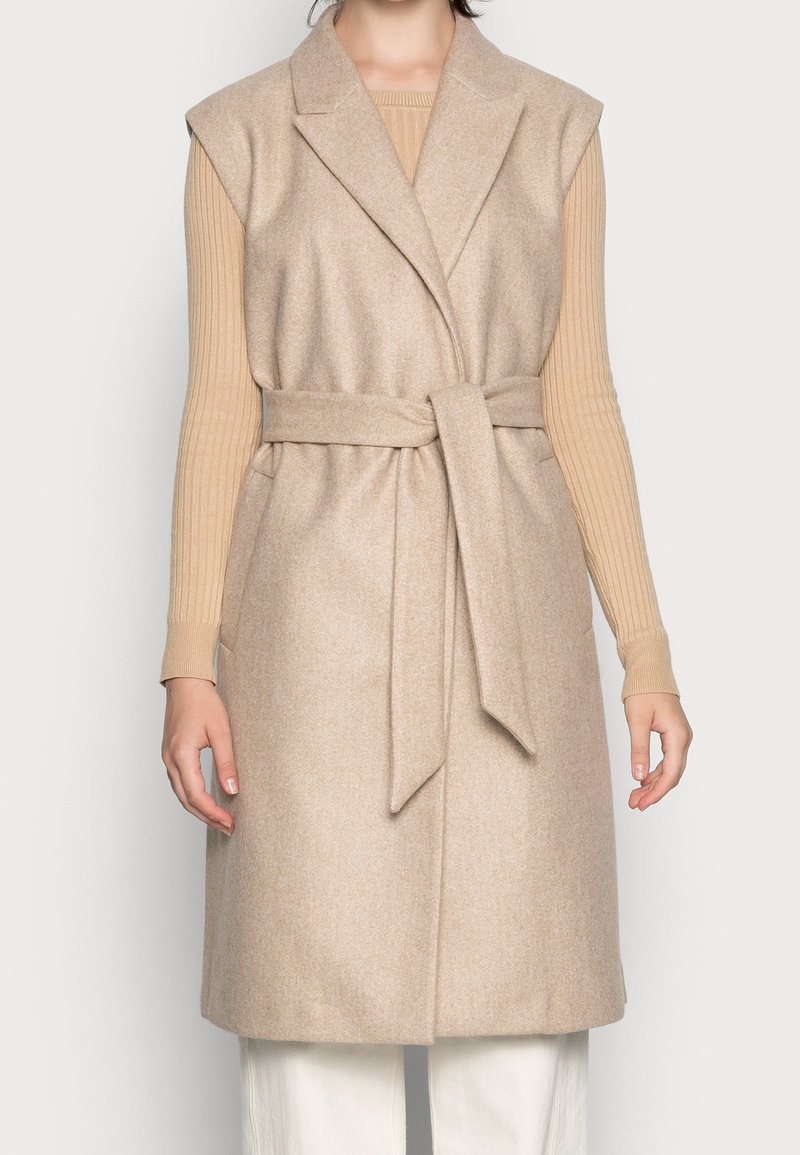 Gilet sans manches en laine beige avec larges revers et ceinture à nouer porté sur un top côtelé à manches longues couleur fauve et un pantalon blanc.