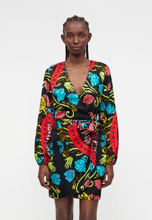 Femme aux cheveux tressés portant une robe portefeuille noire avec un motif floral et de mains coloré, manches longues et un nœud à la taille.