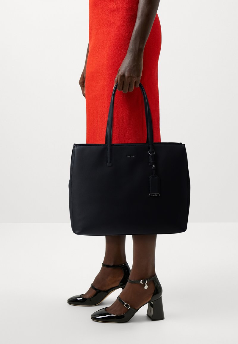 Borsa tote in pelle nera con due manici, forma rettangolare, logo frontale, etichetta coordinata; indossata con un vestito rosso e tacchi neri.