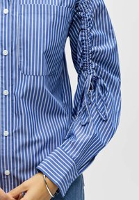 Chemise boutonnée bleu et blanc à rayures avec une poche poitrine, un détail de manches froncées, et un tissu en coton lisse. Accentuée par des boutons blancs.