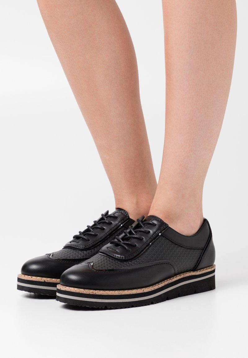 Chaussures en cuir noir texturé à lacets, semelles épaisses rayées, portées pieds nus sur fond blanc.