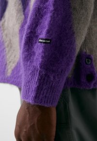 Suéter de lana en tonos morado y gris con un acabado texturizado, que presenta una etiqueta en la manga y botones decorativos en la parte delantera.