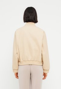 Μπεζ πουλόβερ με μαλακή υφή, ψηλό γιακά, ριμπ μανσέτες και ζώνη μέσης. Η πίσω όψη δείχνει άνετη εφαρμογή και μίνιμαλ σχεδιασμό.