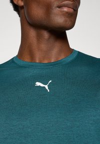 Tyrkisen treningstopp laget av glatt stoff, med rund halsutskjæring og en hvit Puma-logo på venstre brystområde.