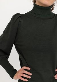 Dunkelgrüner Rollkragenpullover aus strukturiertem Stoff, mit puffartigen Ärmeln und gerippten Bündchen, der eine figurbetonte Silhouette bietet.