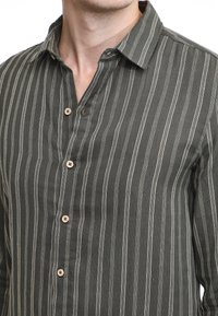 Camicetta button-up verde scuro con strisce bianche verticali, colletto e bottoni in legno. Il tessuto appare testurizzato con un design casual.