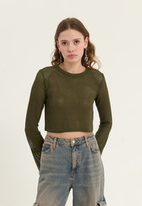 Olivgrön långärmad mesh-crop top med rund hals och intrikat öppet stickmönster, kombinerad med ljusa denimjeans.