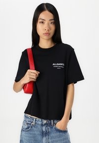Camiseta de algodón negra con cuello redondo y mangas cortas, que presenta un texto de logo en blanco. Combinada con unos jeans de mezclilla azul y un bolso cruzado rojo.