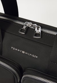 Crna platnena torba s dva srebrna munja, sjajnim metalnim detaljima, dva prednja džepa i utisnutim logotipom: "TOMMY HILFIGER."