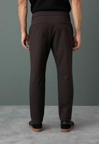 Pantaloni tapered marrone scuro con tasche posteriori, texture sottile e finitura liscia, indossati con scarpe nere su uno sfondo grigio.