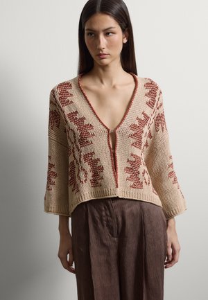 Mujer con suéter beige de punto con patrones geométricos en color óxido y escote en V profundo, combinado con pantalones marrones a rayas.