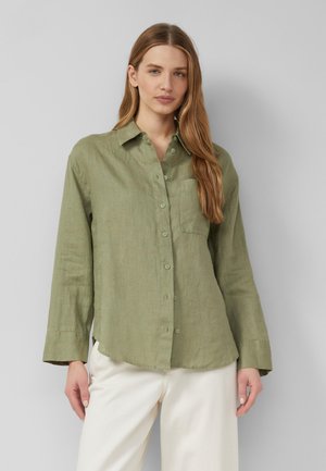 Junge Frau mit langen Haaren, die ein lockeres olivgrünes Hemd mit Knöpfen und weiße Hose trägt, stehend vor einem schlichten grauen Hintergrund.