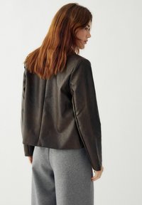 Barna bőrkabát egyenes szabással és sima textúrával, szürke melegítőnadrággal párosítva, hátulról nézve.