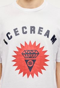 Λευκό βαμβακερό μπλουζάκι με έντονη μαύρη επιγραφή "ICECREAM", συνοδευόμενο από κόκκινο αστέρι και διαμαντένιο σχέδιο, που περιλαμβάνει ένα σύμβολο δολαρίου μέσα σε τρίγωνο.