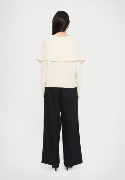 Un pull de couleur crème avec un col replié, associé à un pantalon noir à taille haute et à jambes larges. Texture lisse et coupe décontractée.