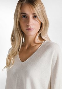 Un pull v-neck beige clair avec une texture douce, présentant une coupe décontractée et de subtils détails côtelés au niveau de l'encolure et de l'ourlet.
