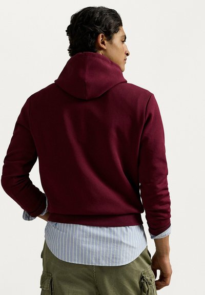 Felpa con cappuccio color melograno dal design aderente. Indossata sopra una camicia a righe azzurro chiaro, presenta un tessuto testurizzato e maniche lunghe.