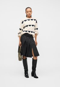 Γυναίκα που φοράει μπεζ πουλόβερ με μαύρες καρδιές, μαύρη πλισέ φούστα με σκίσιμο, μαύρες μπότες μέχρι το γόνατο, κρατώντας μια τσάντα σε ελιά πράσινο χρώμα.