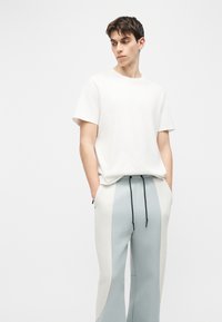 Νεαρός άνδρας με κοντά σκούρα μαλλιά που φοράει απλό λευκό μπλουζάκι και ανοιχτό μπλε και λευκό παντελόνι με κορδόνι, με τα χέρια στις τσέπες.