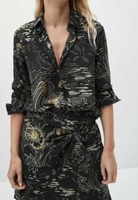 Chemise noire à boutons avec des manches retroussées, ornée d'un motif texturé de fleurs et de vagues en or et en vert. Accompagnée d'une jupe nouée assortie.