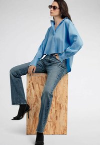 Camicia corta in satin azzurro chiaro con design a bottoni, abbinata a jeans a palazzo blu scuro e scarpe col tacco nere, seduta su un blocco in OSB.