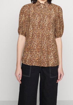 Kvinde iført brun bluse med leopardprint med korte puffede ærmer og afslappede sorte bukser med synlige hvide syninger.