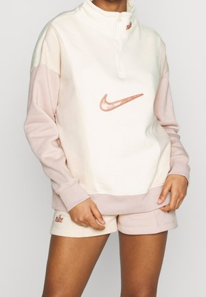 Person trägt ein hellrosa-weißes Nike-Sweatshirt mit Viertelreißverschluss und passende Shorts mit Nike-Logos auf Brust und Bein.