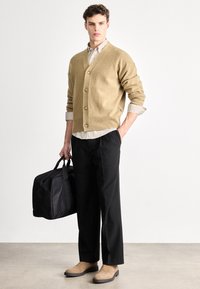 Bēša džempera kardigans ar pogām, svītraina krekla, melnas bikses, gaiši brūnas kurpes un melna duffle soma. Vienkāršs, mūsdienīgs dizains.