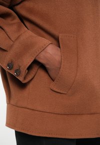 Giacca di lana marrone con una finitura testurizzata, dotata di una tasca laterale e due bottoni scuri sul polsino. Design semplice e dritto.