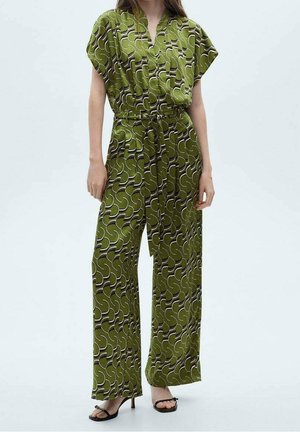 Kobieta ubrana w zielony, wzorzysty kombinezon z krótkimi rękawami, przewiązany paskiem w talii, o szerokich nogawkach, nosząca czarne buty na obcasie z odkrytymi palcami.