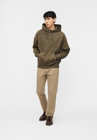 Jeune homme debout les mains dans les poches, portant un sweat à capuche GAP vert olive, un pantalon beige et des chaussures à lacets marron foncé sur fond blanc.