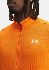 Oranžinė sportinė ketvirčio užtrauktuko viršutinė dalis su tekstūruota geometrine raštu, turinti juodą užtrauktuką ir baltą "Under Armour" logotipą ant krūtinės.