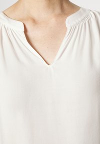 Camicia bianca leggera con scollo a V. Il tessuto è liscio e leggermente strutturato, con sottili arricciature sulle spalle.