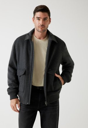 Giacca bomber grigio scuro con zip, due tasche frontali e polsini a coste. Indossata sopra una maglietta chiara e jeans neri.