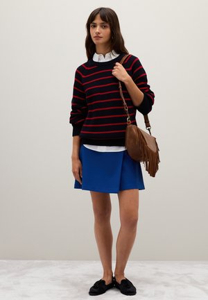Maglione nero a maglia con strisce rosse, camicia bianca con colletto sotto, gonna blu e borsa a tracolla frangiata marrone. Scarpe nere senza lacci.