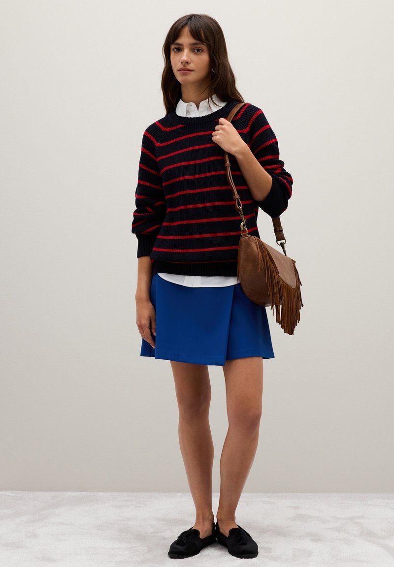 Maglione nero a maglia con strisce rosse, camicia bianca con colletto sotto, gonna blu e borsa a tracolla frangiata marrone. Scarpe nere senza lacci.