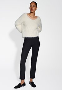 Cremfarbener, kuscheliger Pullover mit weitem Rundhalsausschnitt, kombiniert mit enganliegenden schwarzen Hosen. Schwarze Loafers runden das Outfit ab und betonen eine elegante Silhouette.