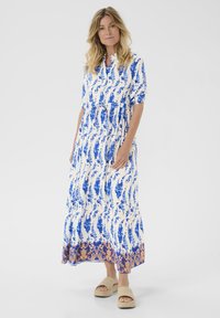 Maxi sukienka z białej tkaniny w niebieski kwiatowy wzór, krótkie rękawy, wcięcie w talii oraz kontrastowy niebiesko-złoty dół. Noszona z beżowymi sandałami.