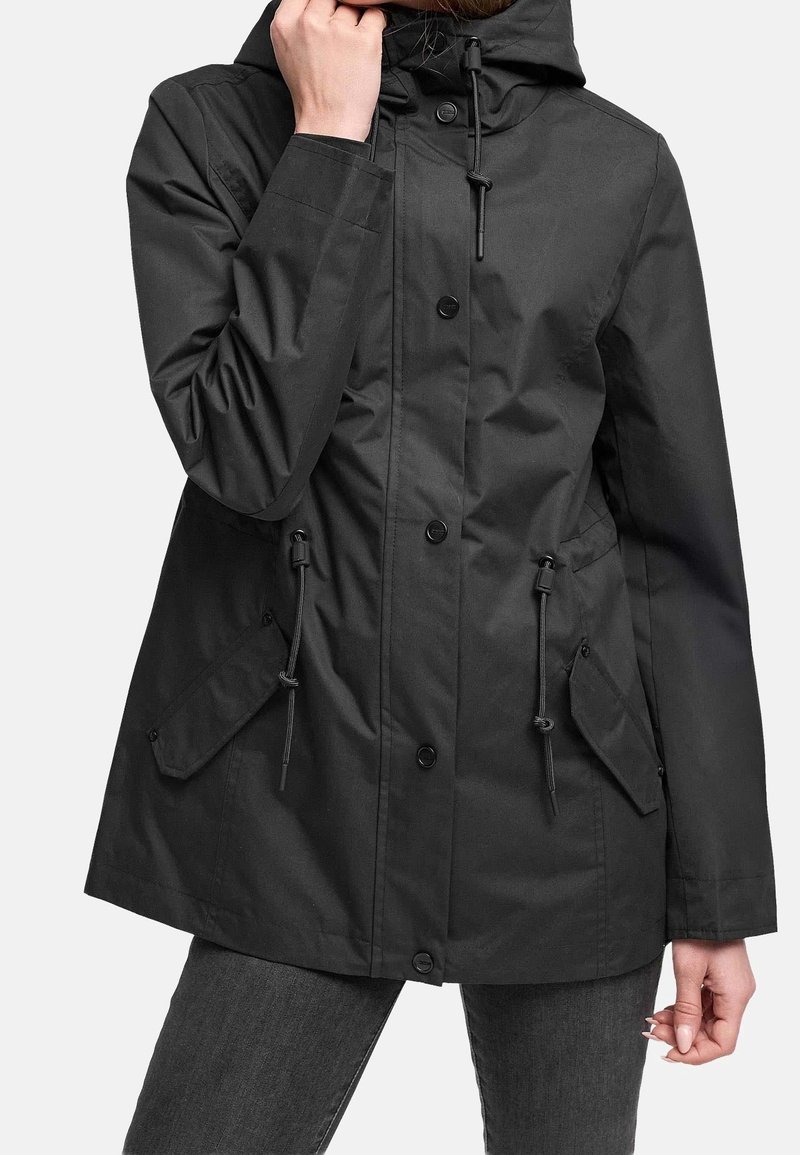 Personne portant une veste à capuche noire avec fermeture par boutons et cordons ajustables à la taille et à la capuche, associée à un pantalon foncé.