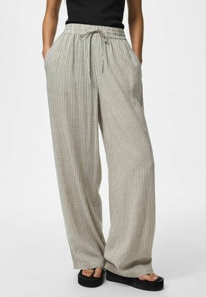 Femme portant un pantalon beige à jambes larges avec motif quadrillé noir, taille élastique avec cordon, haut noir et sandales compensées noires.