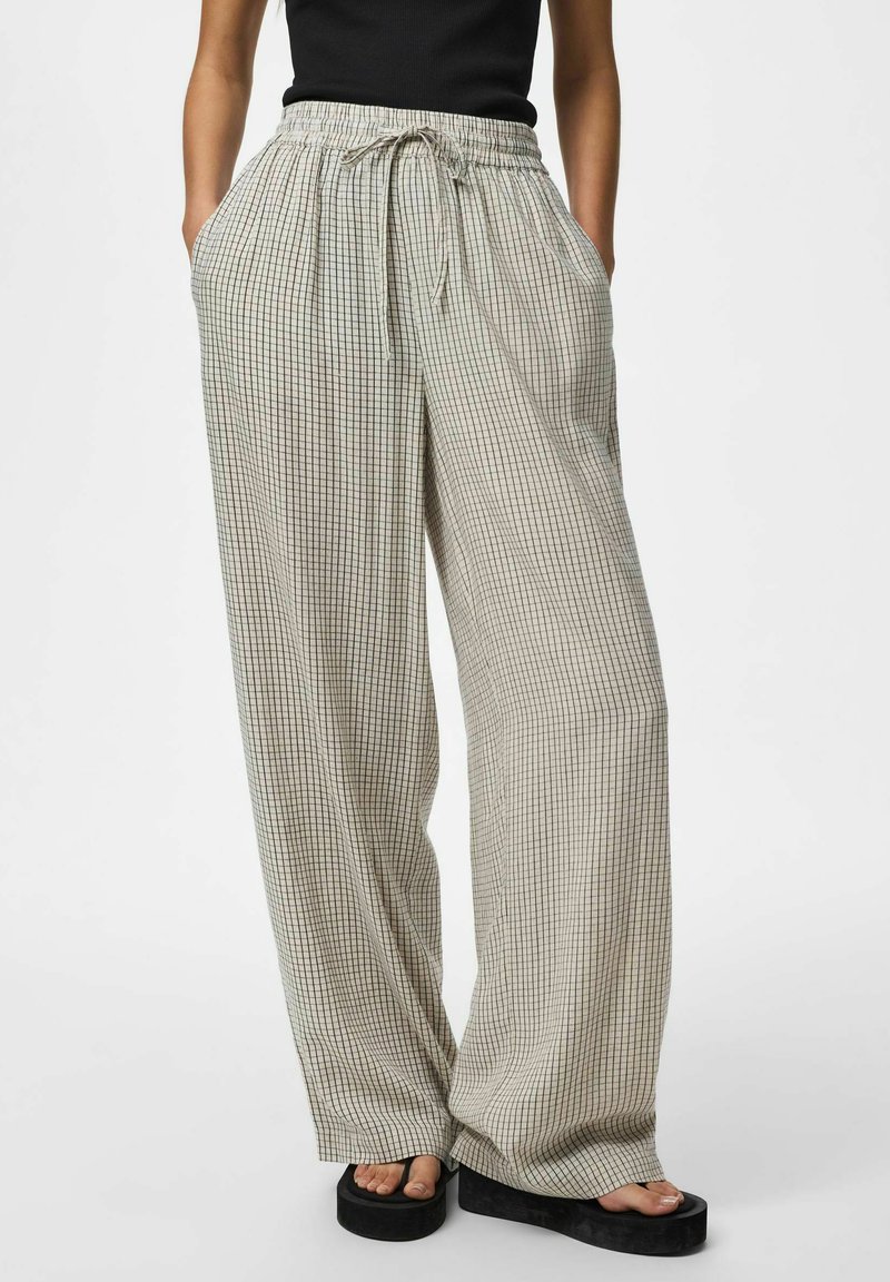 Žena nosiaca béžové široké nohavice s čiernym mriežkovým vzorom, elastický pás so šnúrkou, čierny top a čierne platformové sandále.