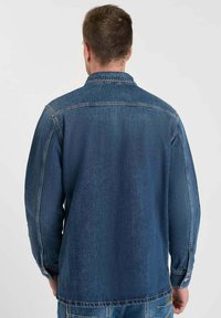 Donkerblauwe denimjas met een klassieke kraag, lange mouwen en een rechte snit. Voorzien van contrasterende stiksels en een zak op de taille.
