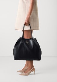 Sort læder tote taske med en tekstureret overflade, to hanke og et trekantet logo. Båret med en ternet nederdel og beige hæle.