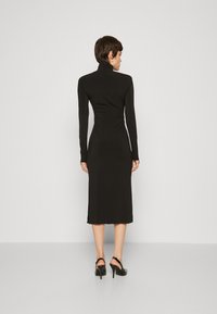 Robe noire à manches longues avec un col montant, design ajusté, longueur mi-mollet, détails froncés à la taille et fentes latérales. Portée avec des talons noirs.