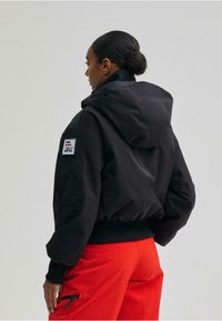 Fekete kapucnis dzseki bordázott mandzsettákkal és szegéllyel, oldalán zsebével és logó foltjával. A modell piros nadrágot visel a dzseki alatt.