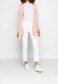 Světle růžový oversized kardigan vyrobený z pleteniny, nošený přes bílý tričko a kombinovaný s bílými zkrácenými džíny a bílými teniskami.