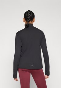 Maglietta sportiva nera a maniche lunghe con collo alto, vestibilità aderente e texture morbida e liscia, abbinata a leggings color bordeaux.
