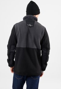 Giacca in pile nera e grigia, con colletto alto, una patch logo sul retro e pannelli in tessuto a contrasto sulle maniche.