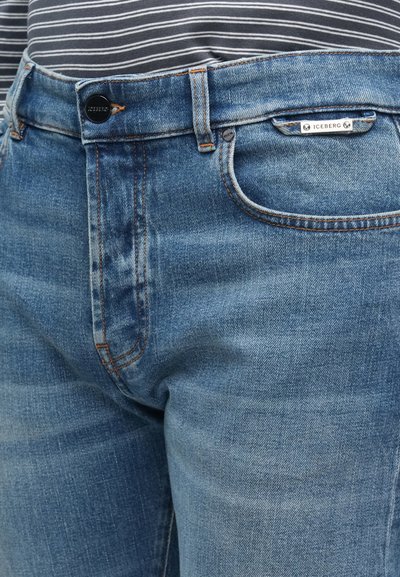 Ανοιχτό μπλε τζιν με ίσια γραμμή, διακοσμημένο με πορτοκαλί ραφές και μεταλλικά εξαρτήματα. Μικρή ετικέτα στη μέση γράφει "ICEBERG".
