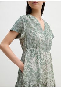 Femme portant une robe courte à fleurs verte et blanche avec un col en V et la main dans la poche, sur un fond uni.
